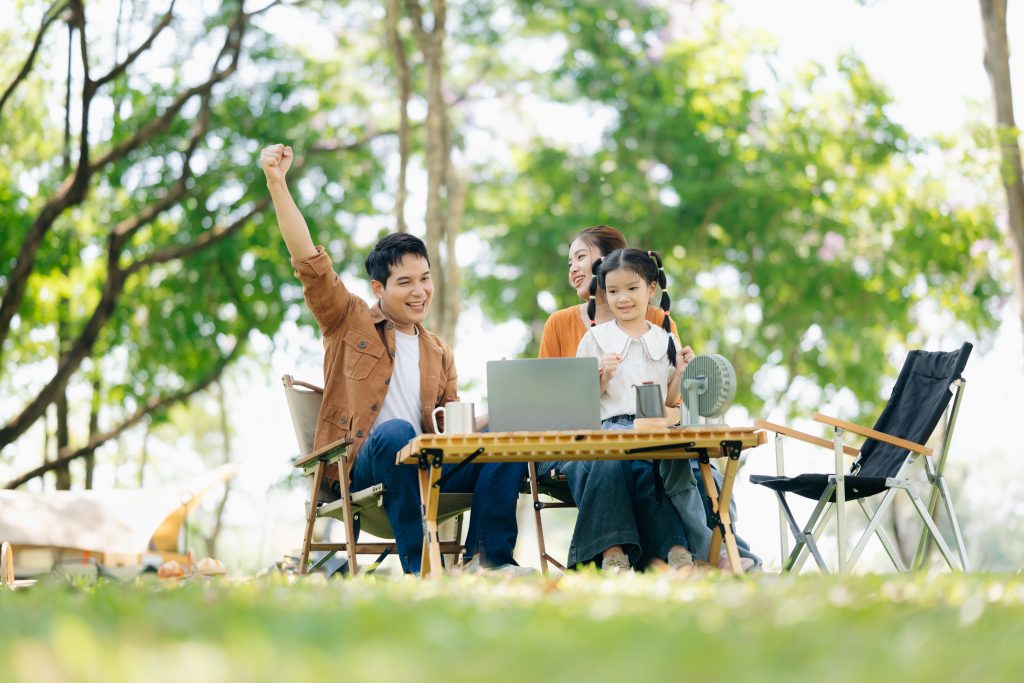 Joyful Asian family camping in a sunny garden, bonding over BBQ, tech, and laughter. Parents and daughter enjoy outdoor love, food, and fun in green summer nature.