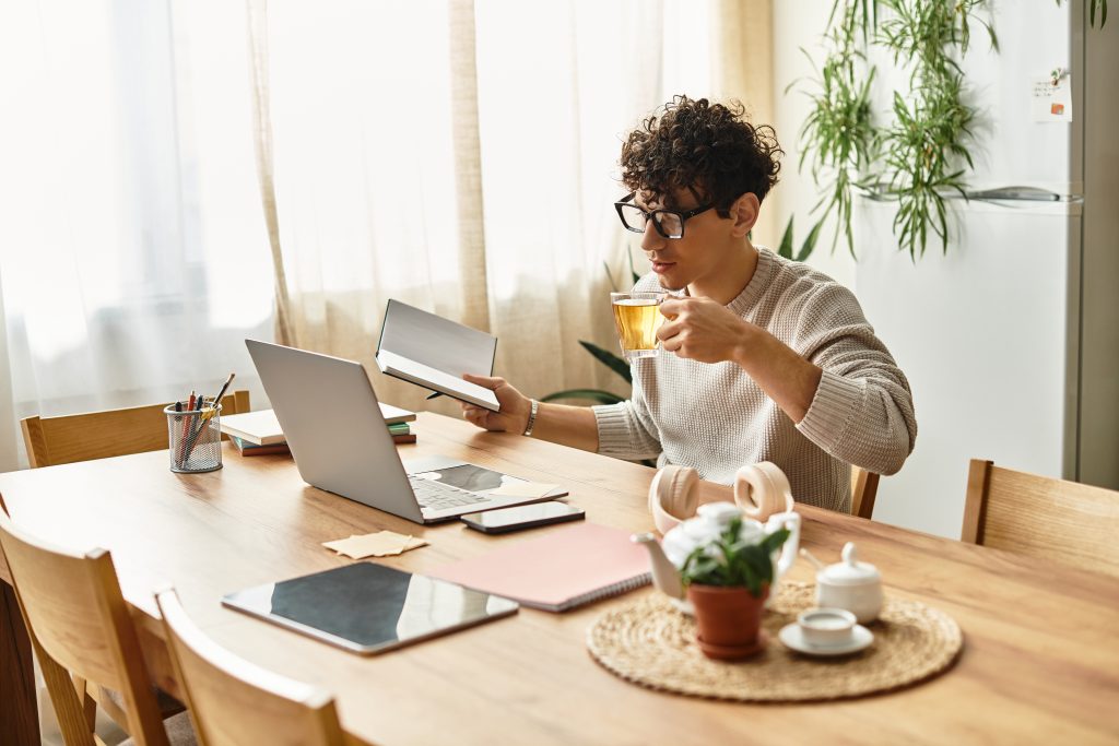 A handsome man sits at a wooden table, sipping tea and reading a book beside a laptop.