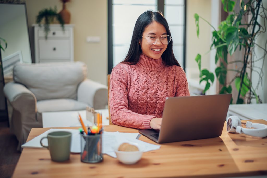 Young Vietnamese woman working in a home office and using a laptop. Flexible workspace and working time. Beautiful Asian business female or student typing an email or working on a project. Copy space.