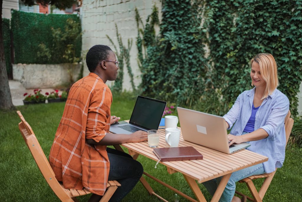Two young businesswomen are sitting at a wooden table in a garden, working remotely using laptops