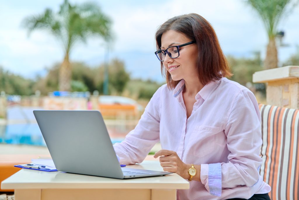 Beautiful business woman in her 40s working on a laptop remotely, sitting at an outdoor table in a resort hotel. Freelance work, business, middle aged people concept