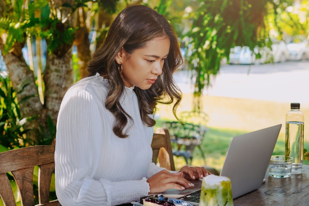 Pretty young student in casual clothing does her homework on laptop outside in sunny day.